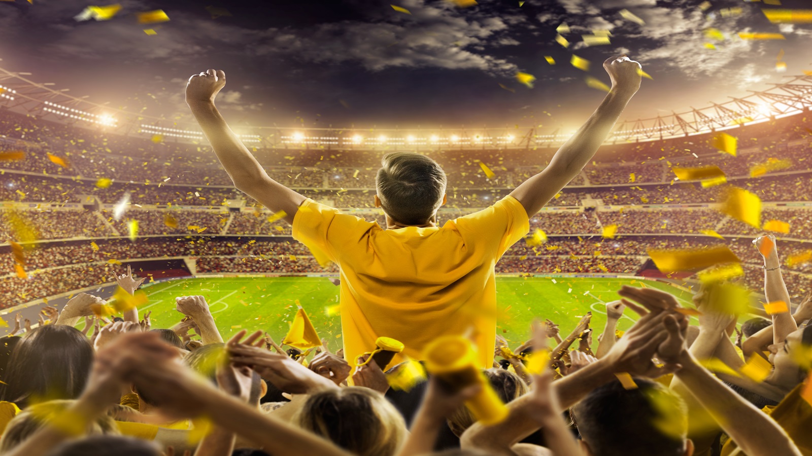 On the foreground a group of cheering fans watch a sport championship on stadium. One man stands with his hands up to the sky. People are dressed in bright colors. A long-range shot of a stadium field, floodlights and seating. A green field, with painted white lines, is visible in the foreground. In the background are diffuse out-of-focus stadium seats. Large, bright floodlights are in the top-left and top-right corners of the image.